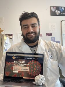 Smiling Amir in a lab coat with a laptop displaying the SciForAll website and a plastic model of a protein