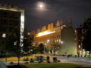 Yellow lights glow from greenhouses on top of the Earth Sciences Building at night, with the moon glowing above and Huron Street below