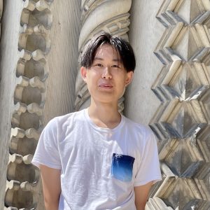 Yutei Shi, an Asian man in a white T-shirt gazing into the camera with carved pillars in the background
