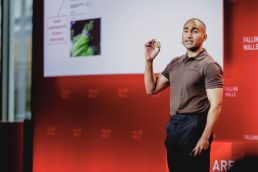 Adnan Sharif stand before a projection screen. He holds up a cube of hydroponic soil, looking at the audience as he explains his project