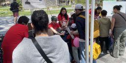 four presenters in red shirts sit under a white tent and show child and adult spectators their activities on folding tables